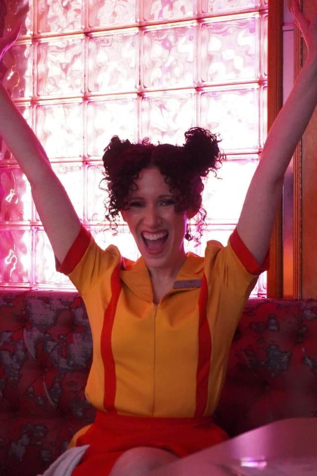 Ali in a yellow diner uniform, arms up against a pink glass-brick wall